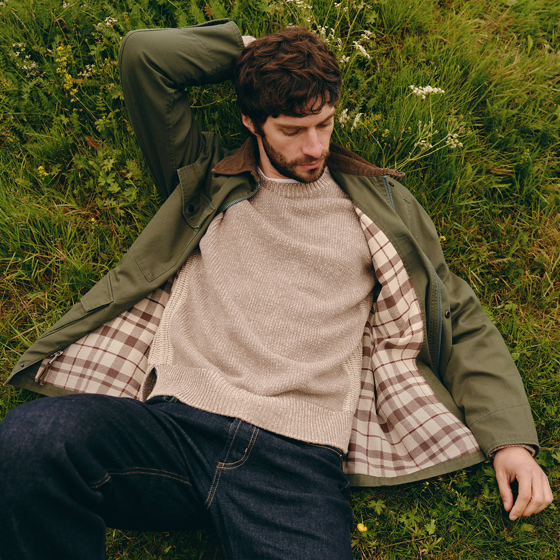 A man lying on green grass wearing an olive green jacket with a brown corduroy collar, a beige knit sweater, a brown and beige plaid shirt, and dark blue jeans.