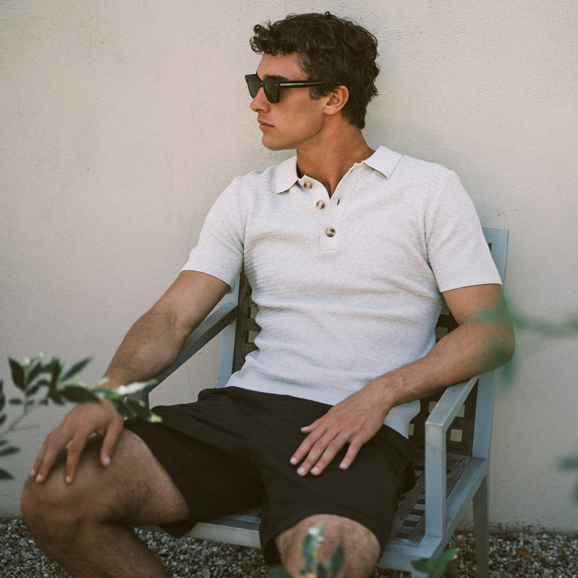 Man sitting on a chair wearing sunglasses and a light-colored polo shirt against a neutral wall.