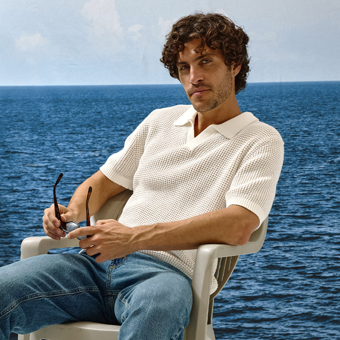 Man sitting on a chair by the ocean wearing a white shirt and blue jeans.