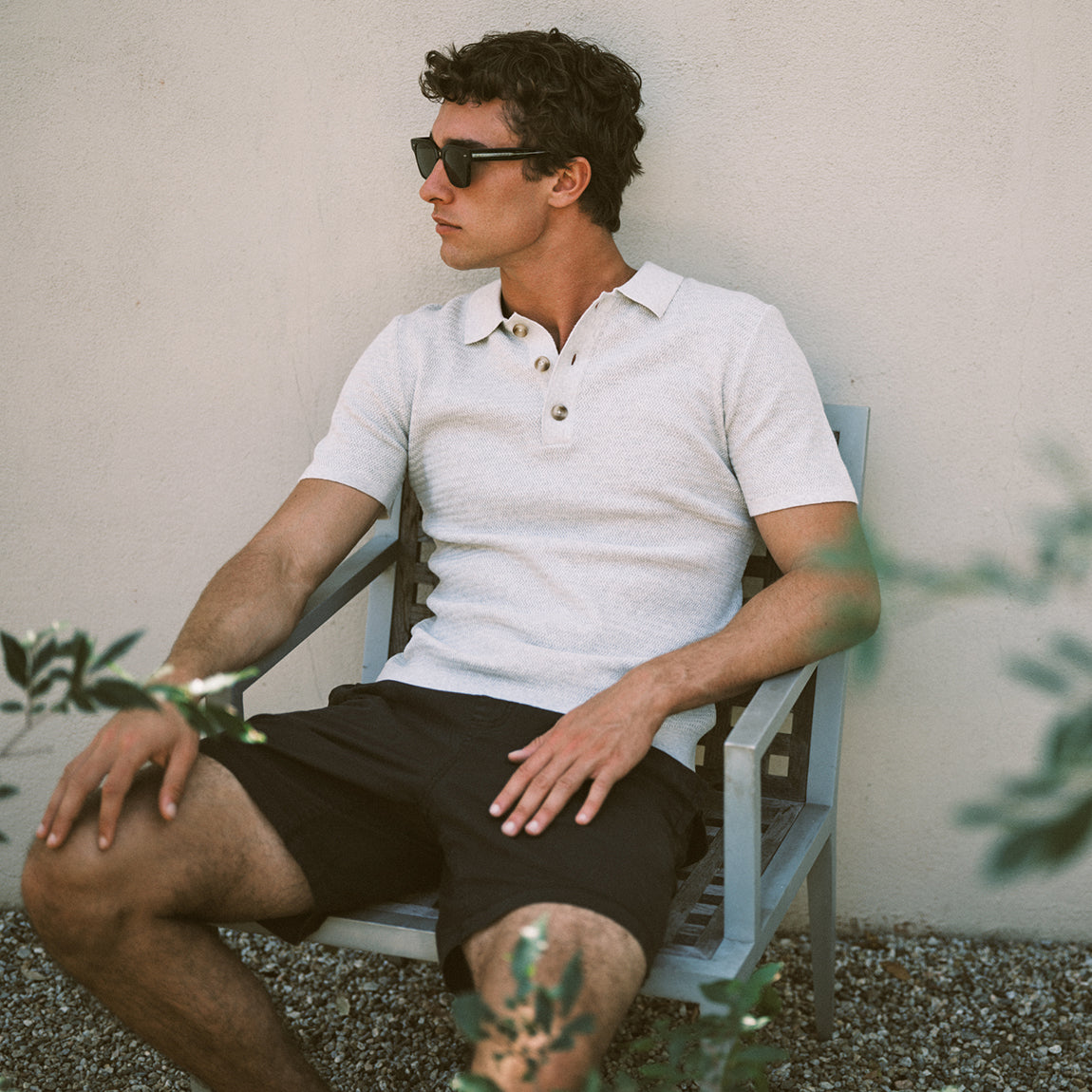A man wearing a white short-sleeve polo shirt and black shorts sits in a light blue wooden chair against a beige stucco wall, wearing dark sunglasses.