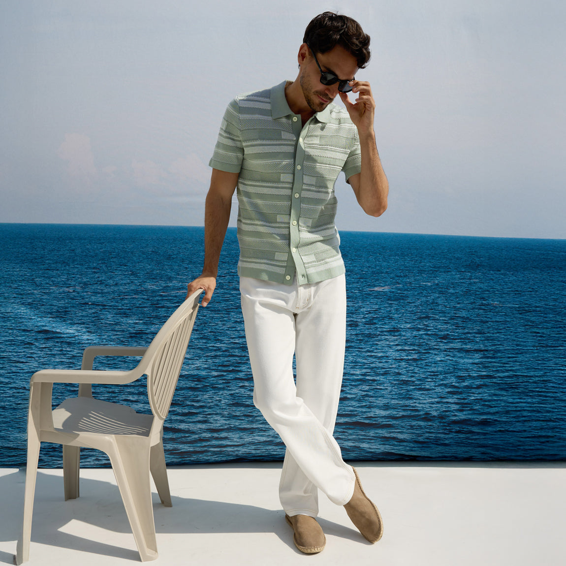 Man in striped shirt and white pants standing by the ocean with a chair.