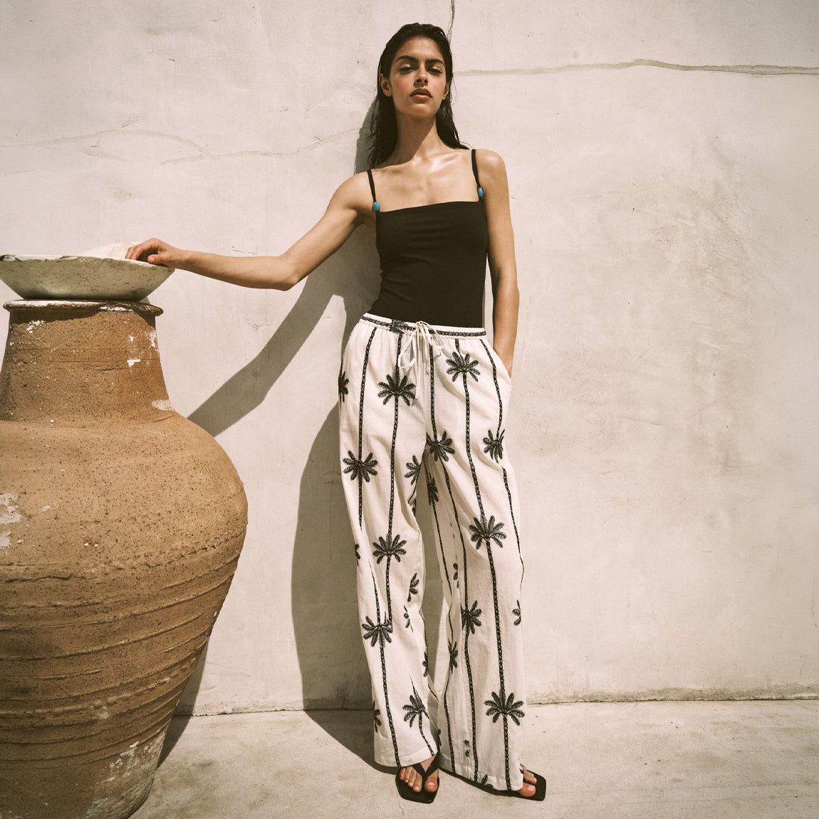 Woman in black top and white pants with black patterns leaning against a textured wall.