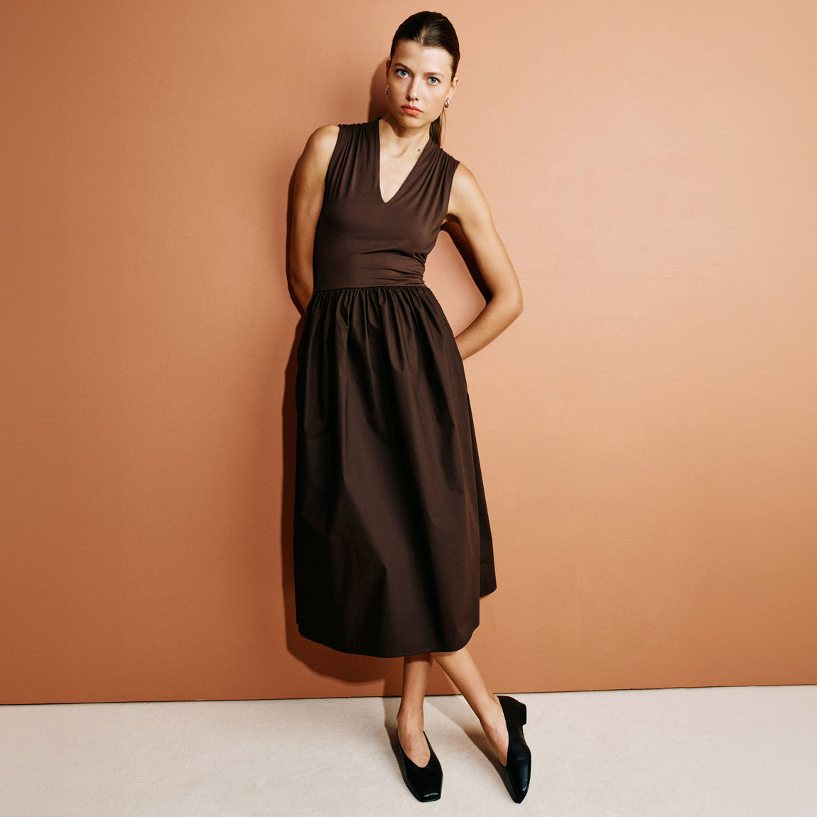 A woman modeling a sleeveless dark brown midi dress with a V-neckline and gathered skirt, paired with black pointed flats, set against a solid terracotta-colored studio wall.