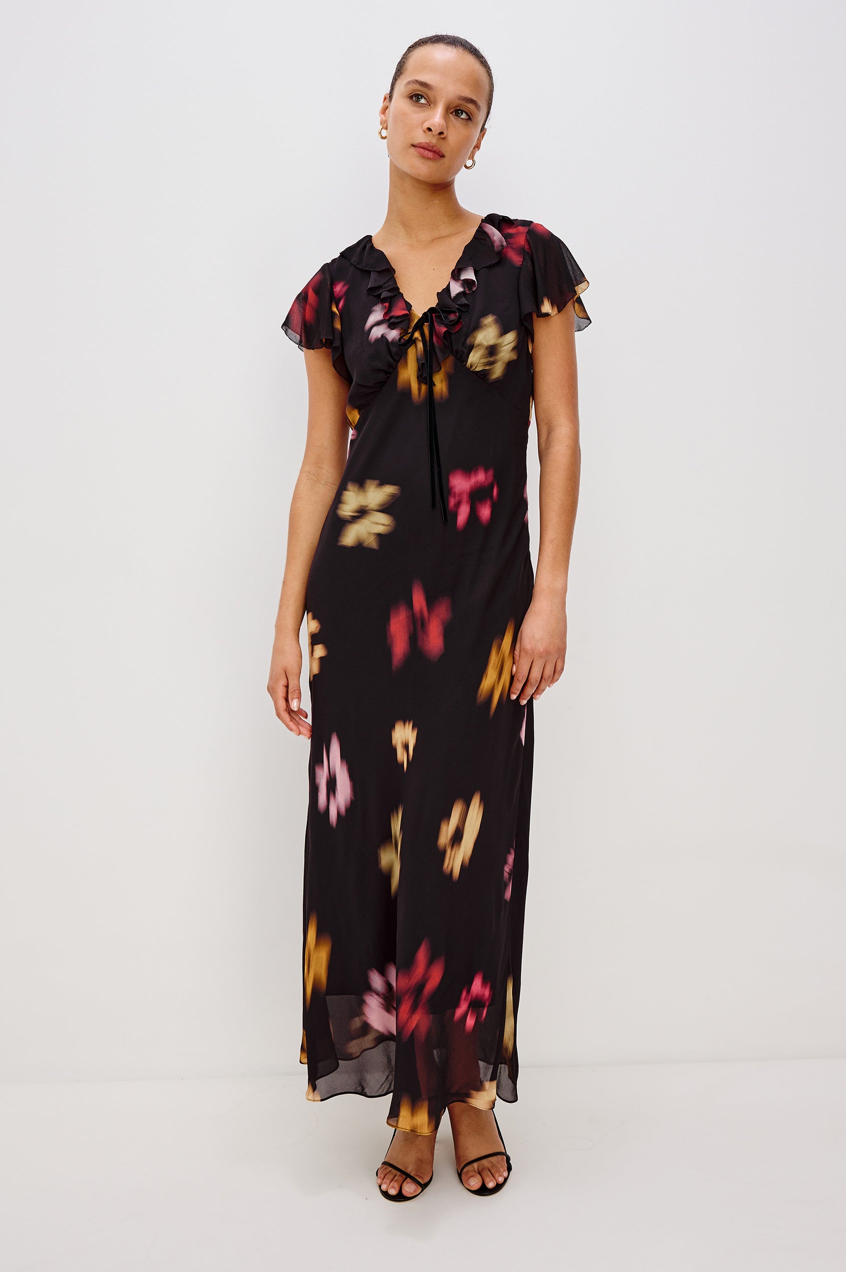 A chiffon midi dress with a blurred floral print. The dress has a V-neckline framed by cascading ruffles and a velvet bow, ruffle cap sleeves, and an empire waist that creates a graceful, elongated silhouette. Full Body Front View featured on Model.