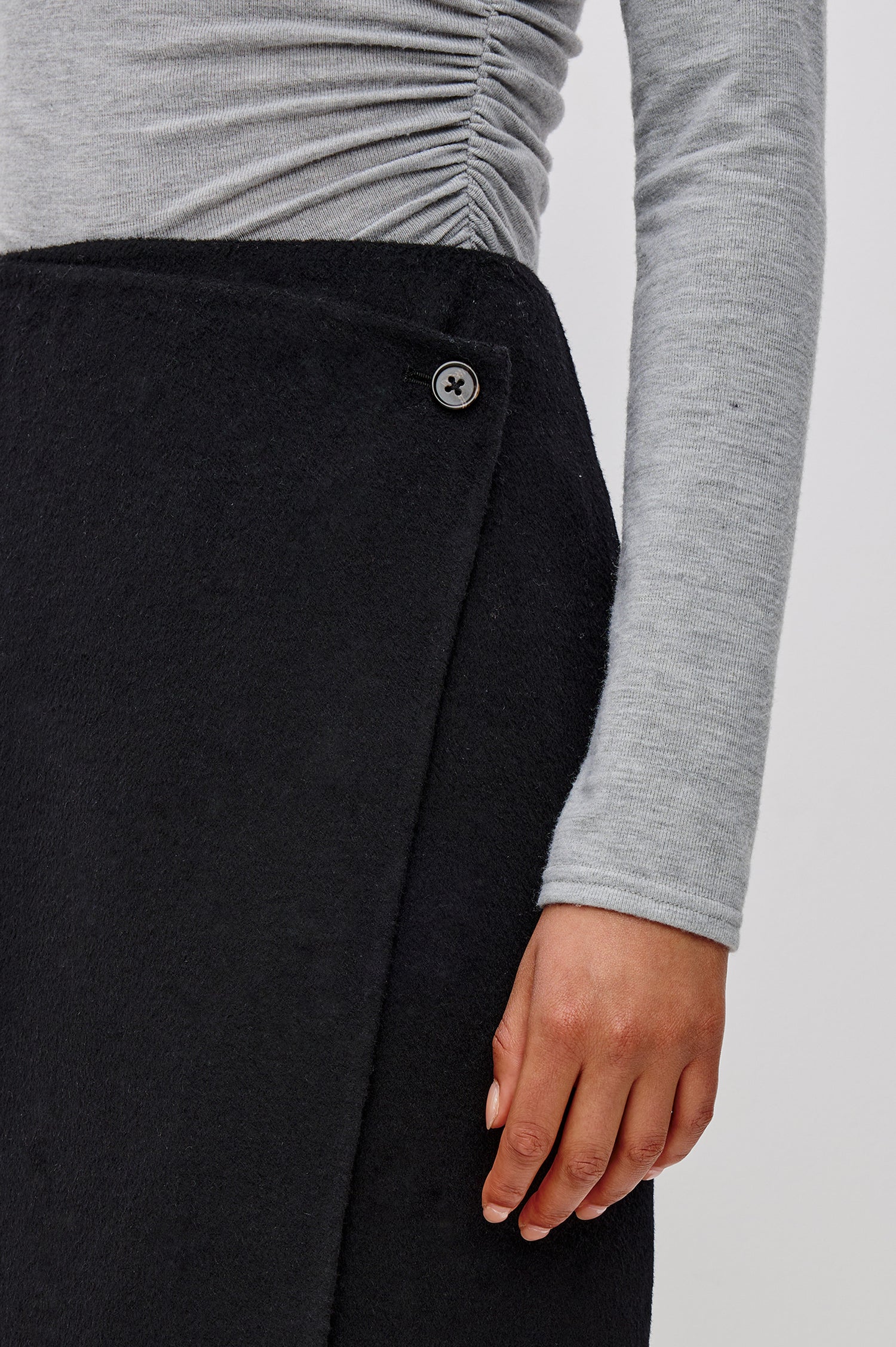 A high-waisted black wool-blend midi skirt. The wrap-style design fastens with a side button and interior closure, finished with statement fringe along the hem for added movement. Close Up View Featured on Model.