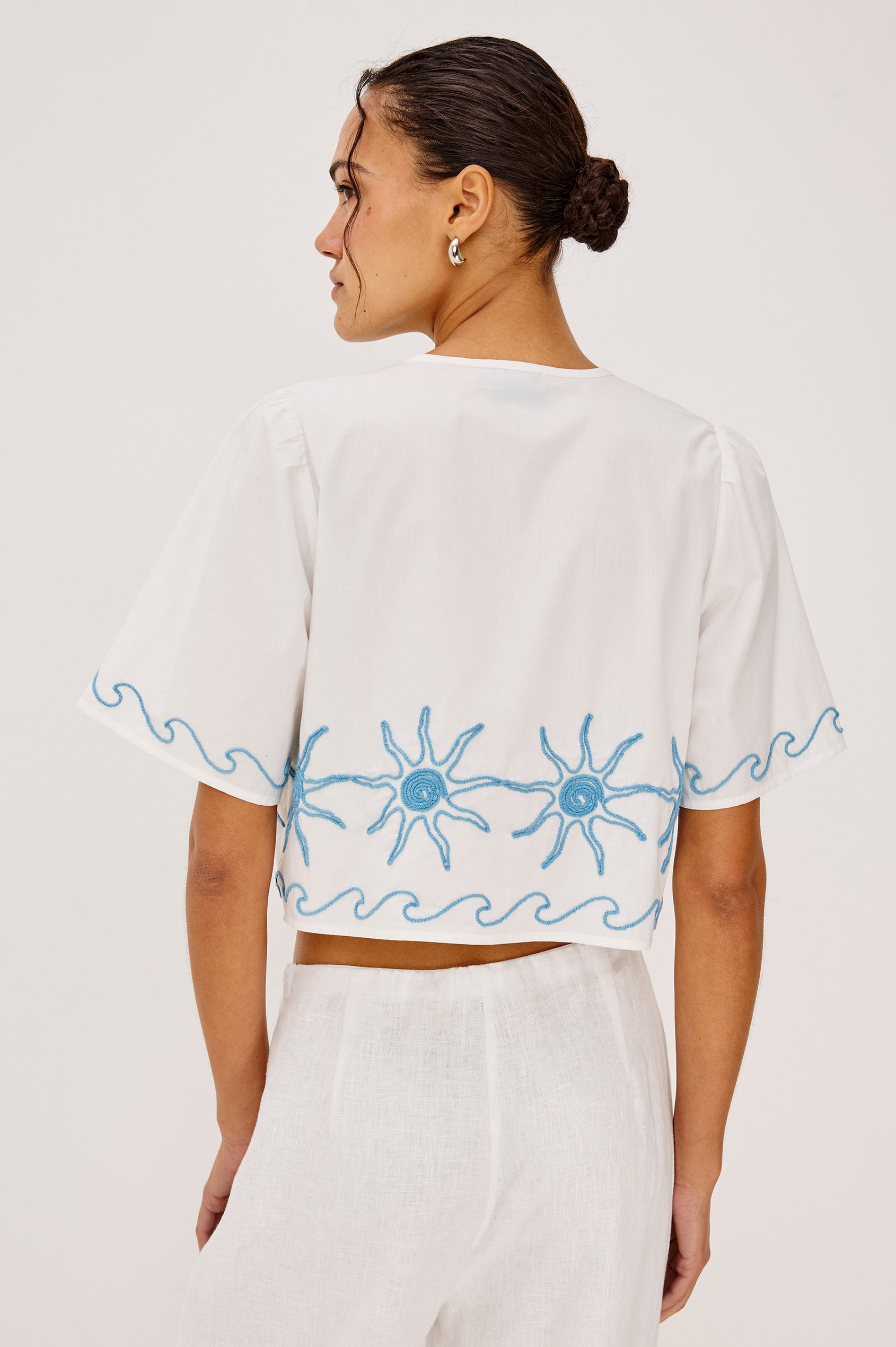 Pari Short-Sleeve Cropped Top with V Neck, Front Tie Closures, and Sun Embroidery Details in Blue Stitching Against White Fabric - Upper Body Back View Featured on Model