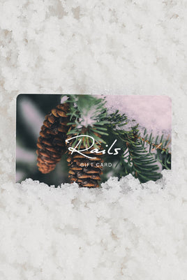 Rails gift card partially tucked into fresh snow, featuring a close-up photo of pine cones hanging from a snow-dusted evergreen branch.