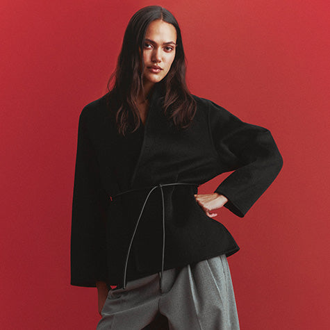 Model with long dark hair in a black wrap jacket tied with a thin leather belt and gray pleated trousers, one hand on her hip, against a red background.