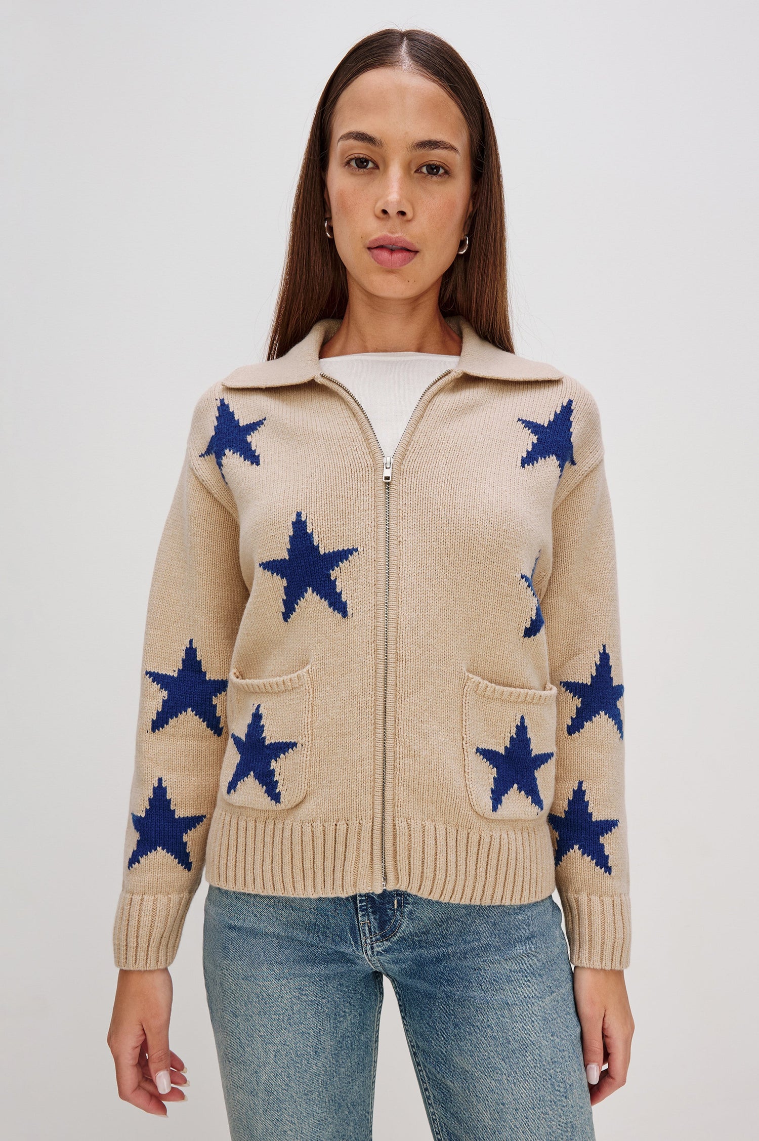 Rhea, a longline camel cardigan in a wool blend, featuring cobalt blue star motifs. The cardigan has a relaxed fit, zip-front closure, and front patch pockets. Front View Featured on Model, Zipped Up