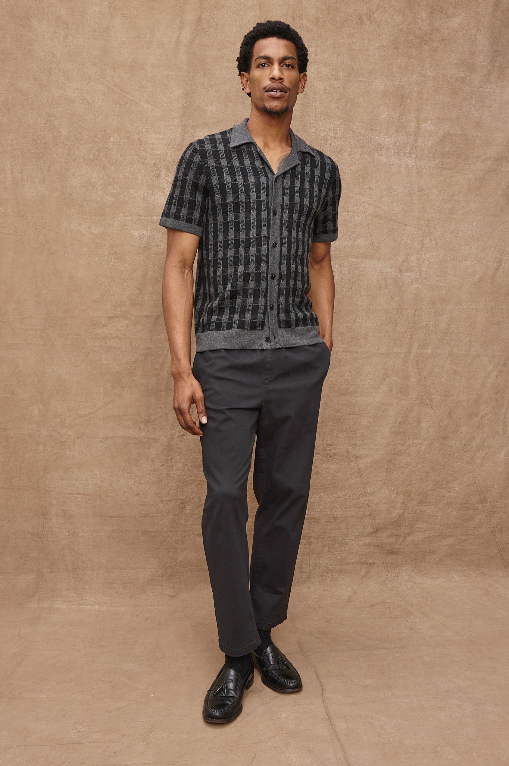 The Ronan is a collared button front knit shirt with short sleeves, grey rib trim, grey + black block stitch pattern in Charcoal Black colorway. Front full body view featured on model.