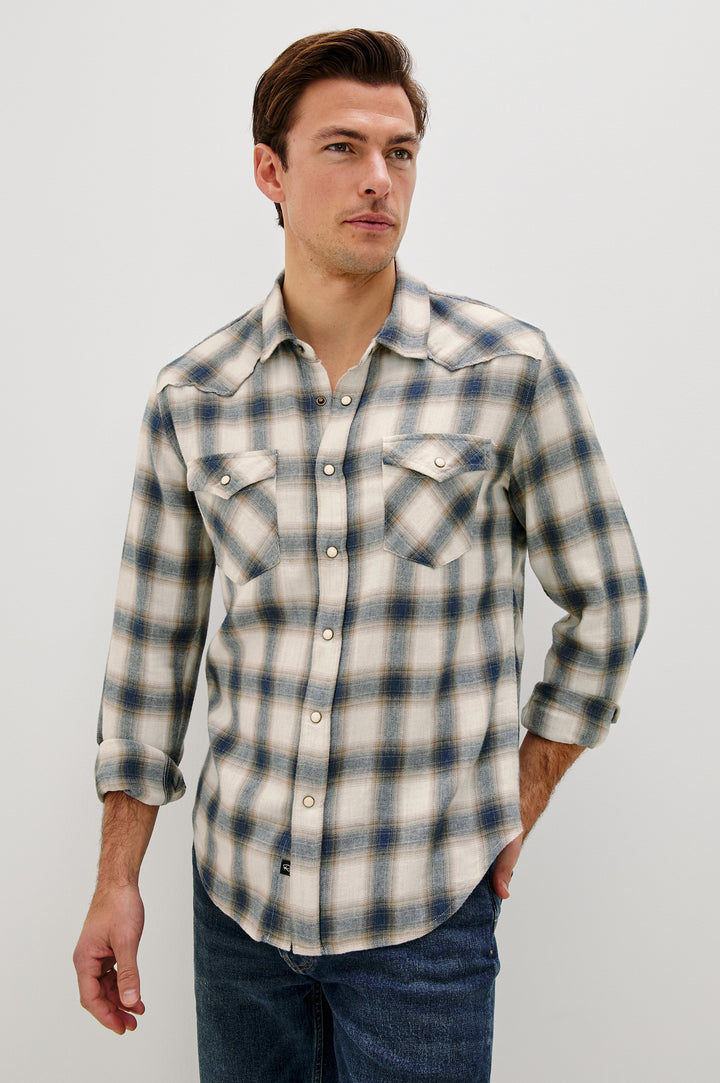 The Roy is a collared snap front shirt with long sleeves, two chest pockets, high low hem and a western yoke seam, in a plaid Wheat Marine Heather colorway. Front view featured on model.