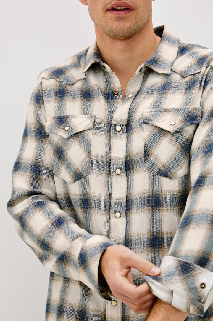 The Roy is a collared snap front shirt with long sleeves, two chest pockets, high low hem and a western yoke seam, in a plaid Wheat Marine Heather colorway. Front detail view featured on model.