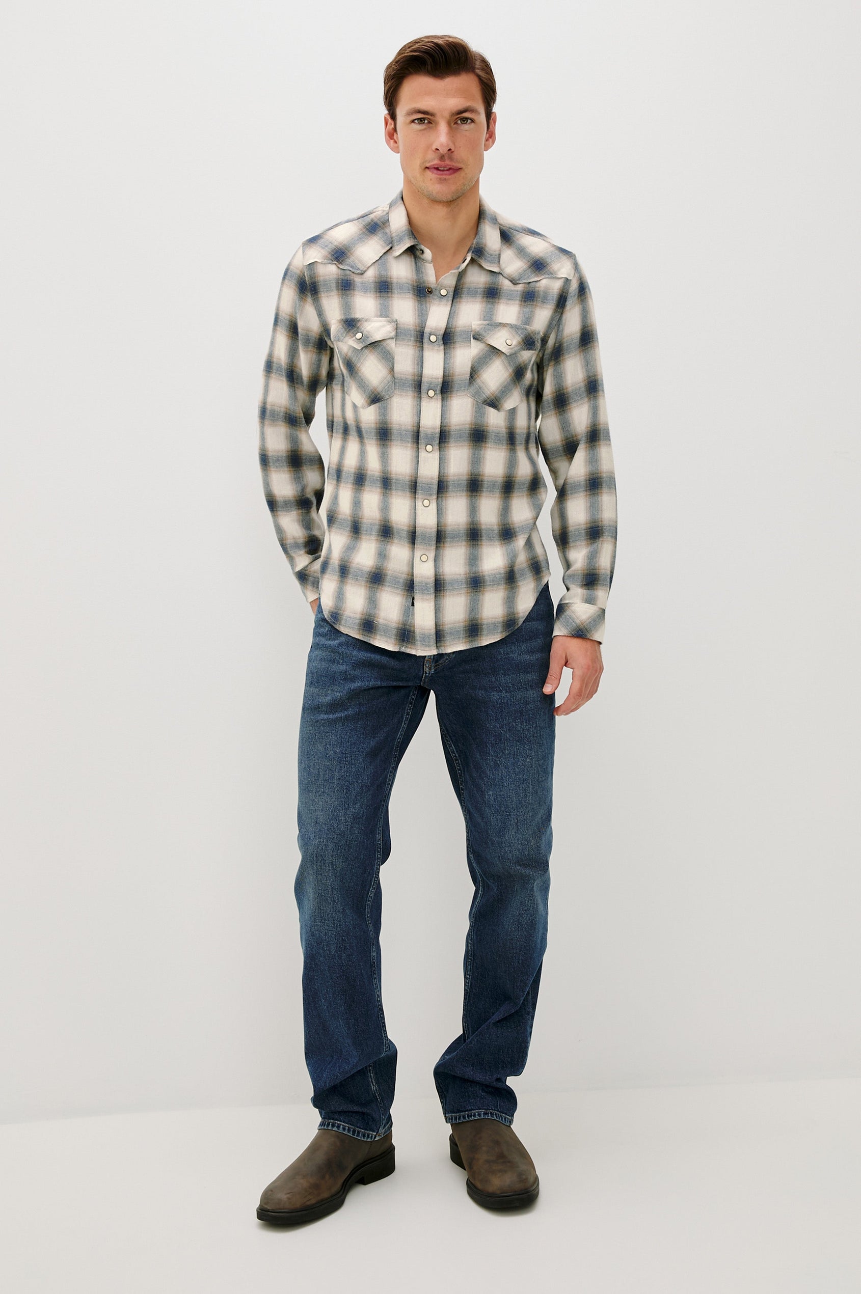 The Roy is a collared snap front shirt with long sleeves, two chest pockets, high low hem and a western yoke seam, in a plaid Wheat Marine Heather colorway. Front full body view featured on model.