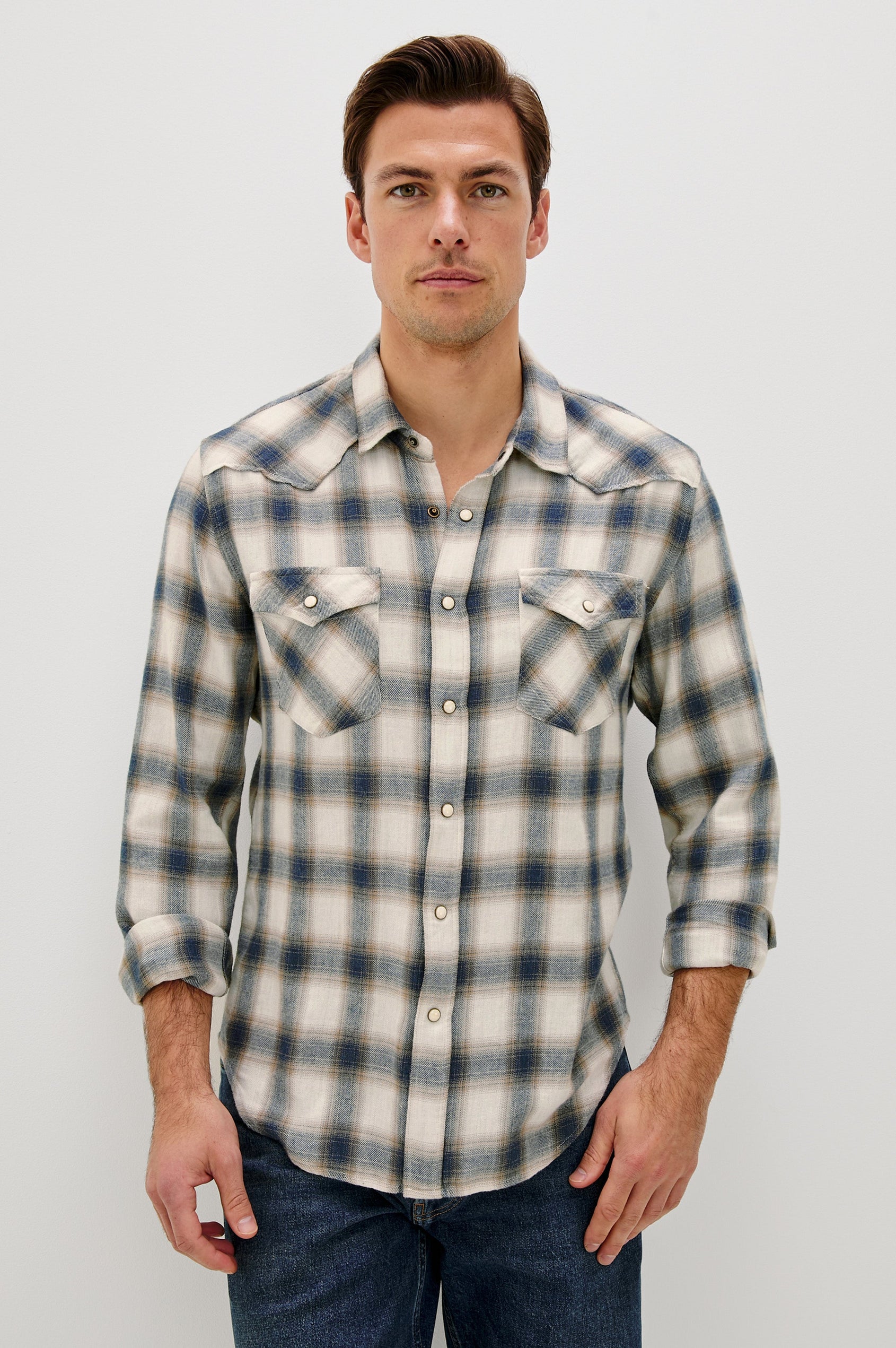 The Roy is a collared snap front shirt with long sleeves, two chest pockets, high low hem and a western yoke seam, in a plaid Wheat Marine Heather colorway. Front view featured on model.