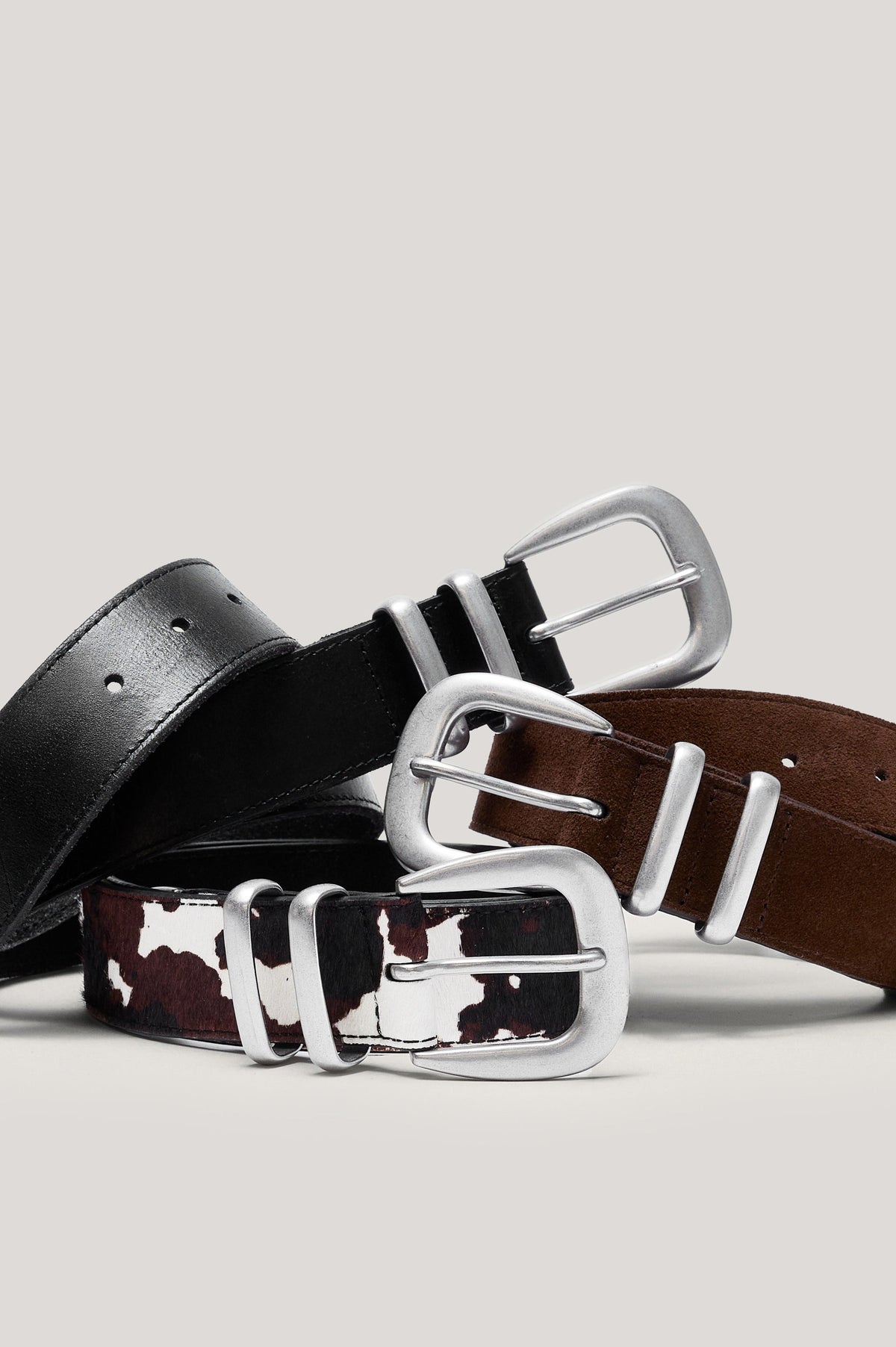 Three colorways of the ryder belt piled together, black leather colorway on top, brown suede leather colorway in the, middle and cow print colorway on bottom.