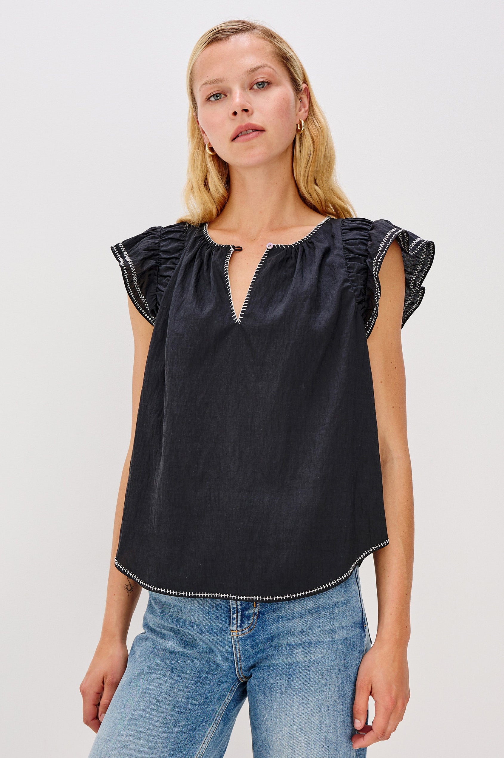 Sable is a scoop neck top with flutter sleeves and a split neck that is adjustable with a button, it has a high low hem and contrast white stitching in Black colorway. Front view featured on model.