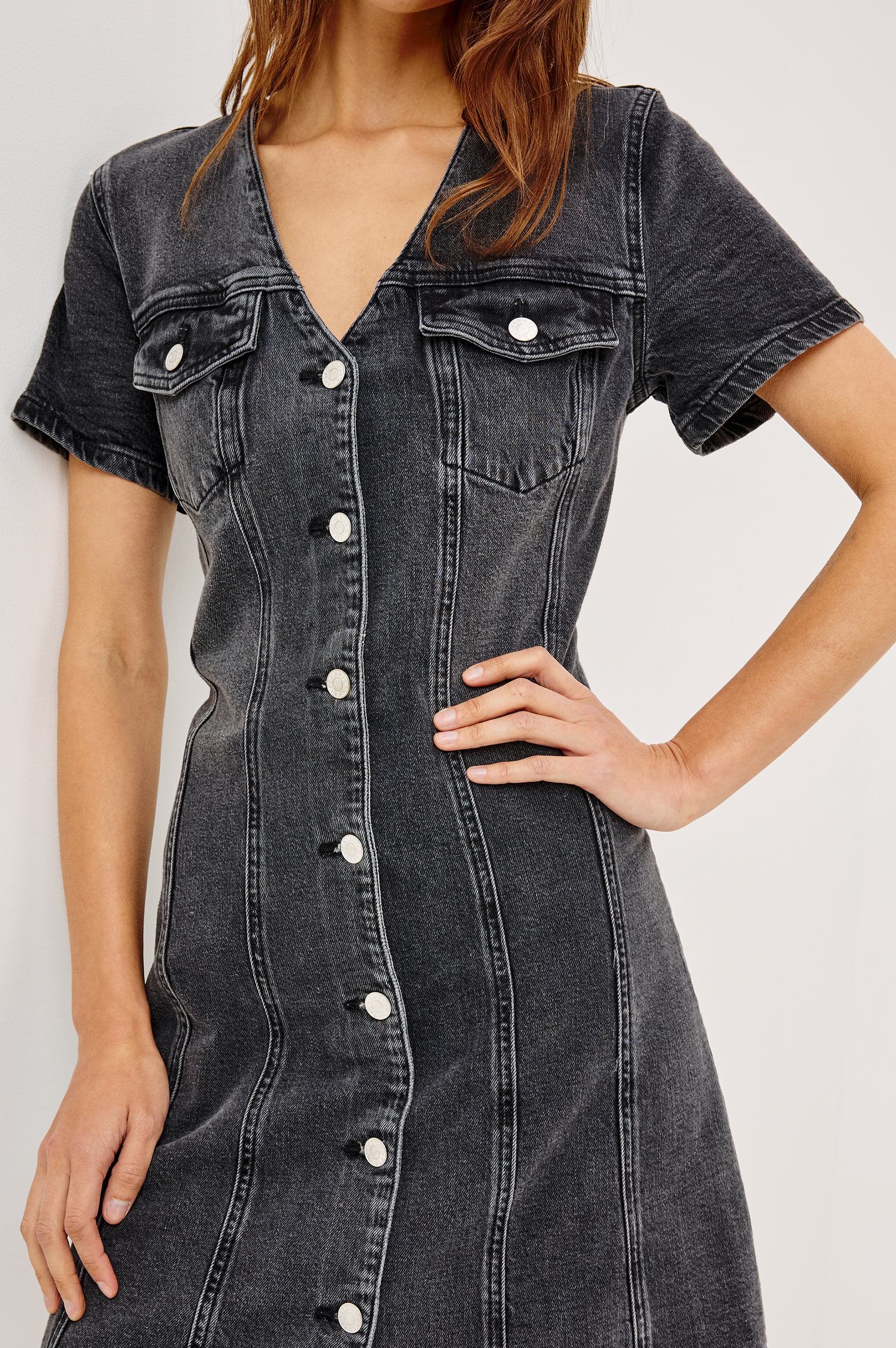 Saratoga Denim Mini Dress With V-Neckline, Buttoned Chest Pockets and Front Buttons, Short Sleeves, Seam Details, and A-Line Silhouette in Washed Black Colorway - Front Close Up View Featured on Model