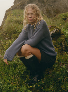 Woman crouching down in a grassy field wearing a blue sweater black skirt and black boots with knee high socks