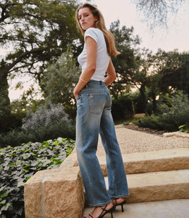 Woman wearing a white baby tee, blue jeans, and black heels while standing in a garden setting.