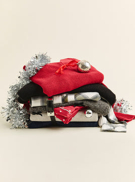 Stack of folded winter clothing, including red and black sweaters, plaid shirts, and a patterned scarf, decorated with silver tinsel and metallic ornaments.