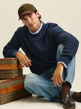 A man wearing a navy blue cable-knit sweater, blue jeans, brown socks, and a brown tassel shoe sits on the floor next to stacked vintage suitcases against a plain beige background.