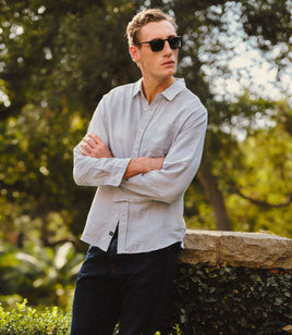 A man wearing a light gray linen button-down shirt and dark sunglasses, standing outdoors with arms crossed against a blurred background of green foliage and a stone wall.