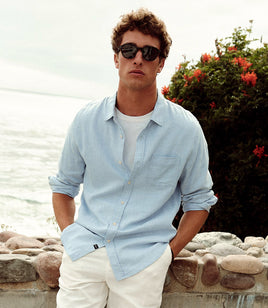 Man wearing sunglasses and a light blue shirt by a body of water with greenery.