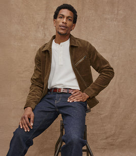 Man wearing a brown jacket and blue jeans sitting on a stool against a beige background