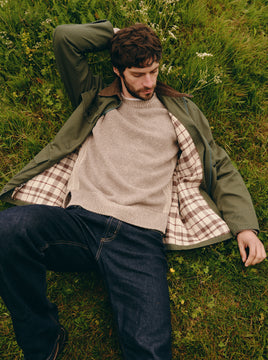 Man wearing a beige sweater with a green overcoat and dark wash jeans while laying in a grassy field.