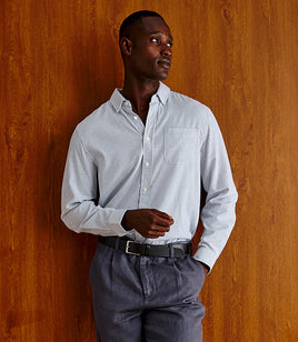 A man wearing a light blue and white vertically striped button-down shirt, gray trousers, and a black belt, standing against a warm wood-paneled wall.