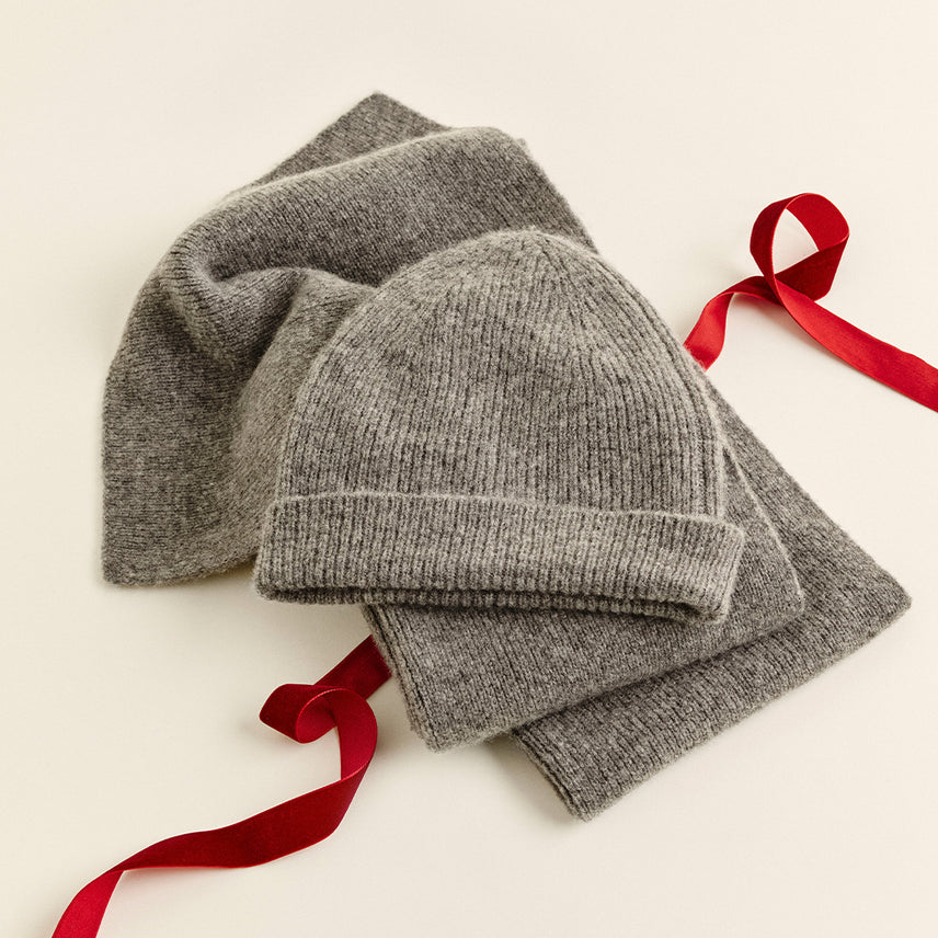 Gray ribbed knit beanie with a folded cuff resting on a matching gray scarf, with a red satin ribbon, on a cream background.