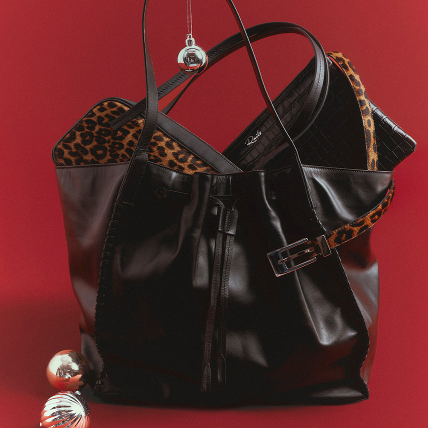 Black leather tote with a drawstring front and whipstitched edges, holding a leopard-print pouch and a black croc-embossed clutch, with a leopard-print belt and silver ornaments, against a red background.
