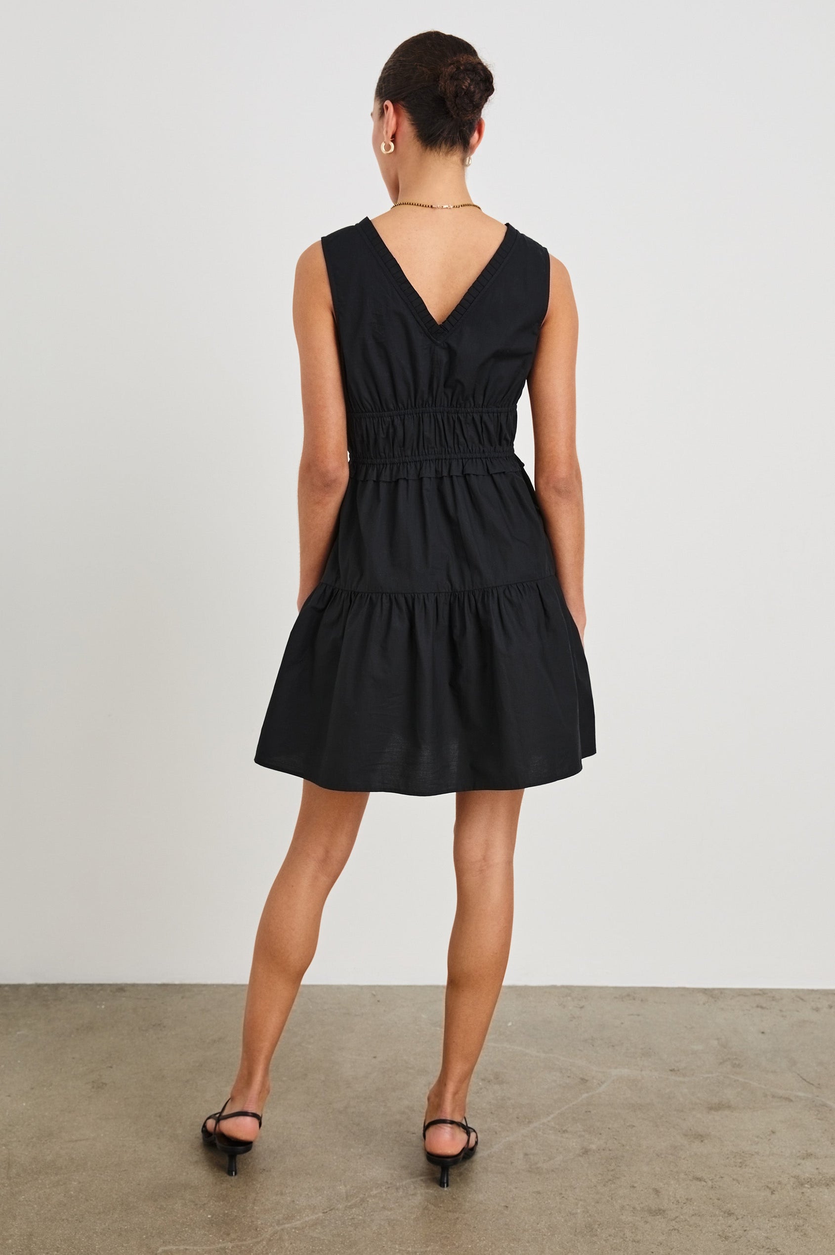 Back, detail view of female model wearing our sleeveless, Talulah Mini Dress in color Black. Dress features pleated v-neck and smocked waist.