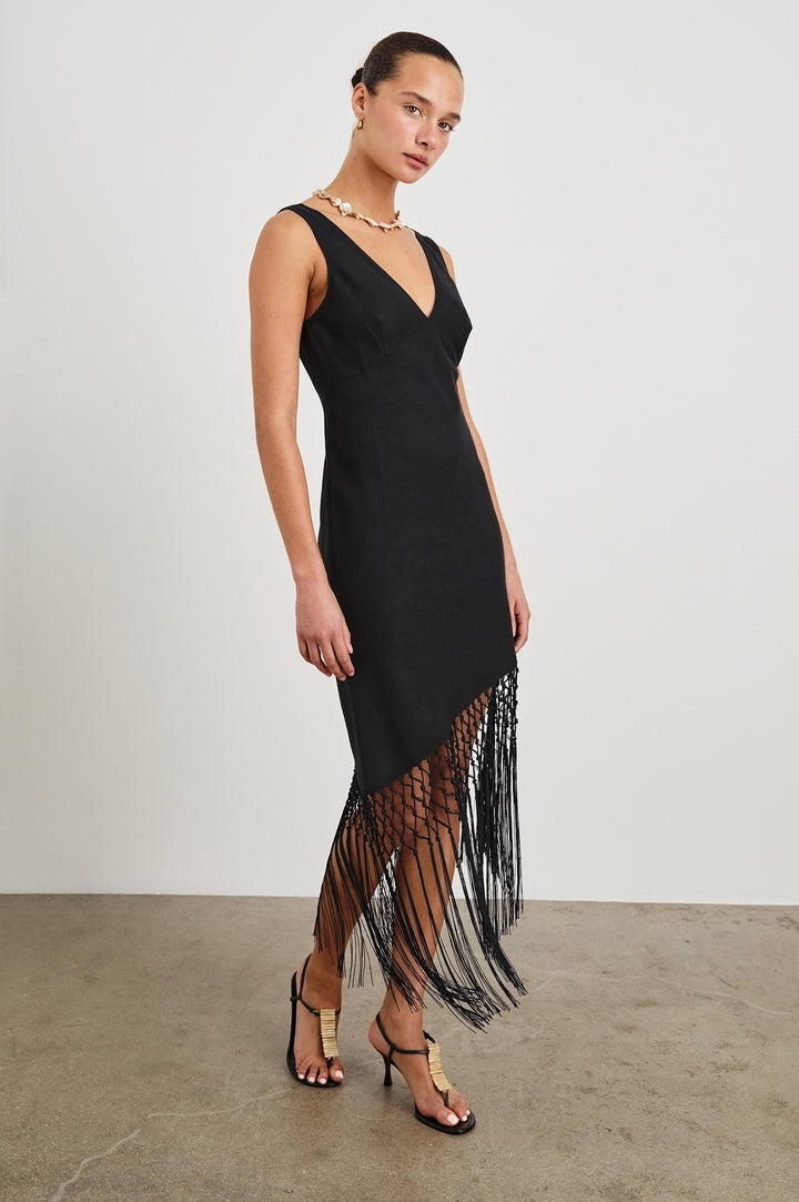 Front view of female model wearing our Tamara Dress in Black.  This is a black mini dress with an asymmetrical fringe hem, sheer netted panel, and empire waistline. 