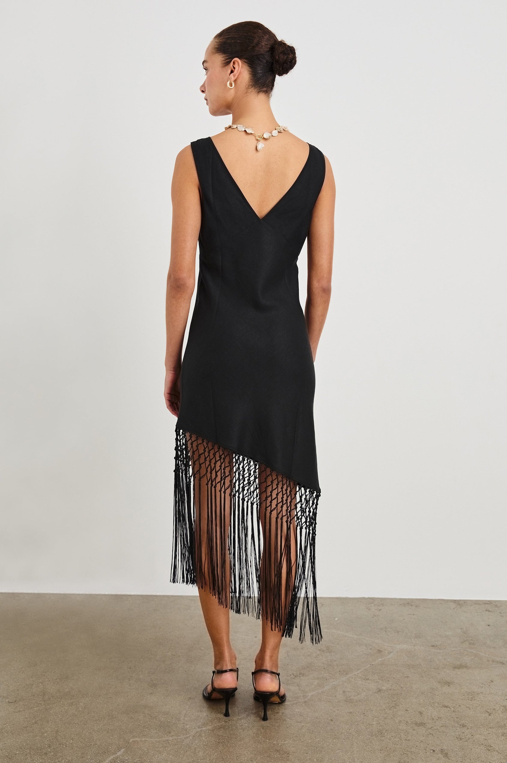 Back view of female model wearing our Tamara Dress in Black.  This is a black mini dress with an asymmetrical fringe hem, sheer netted panel, and empire waistline. 