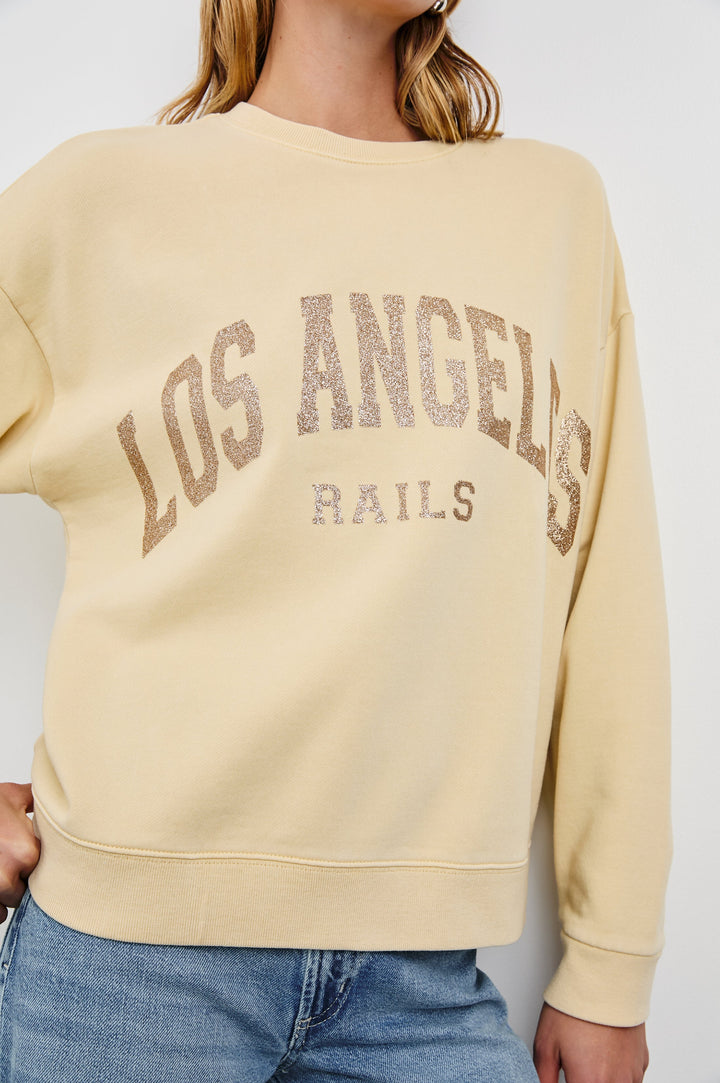 The Varsity Sweatshirt with a Crew Neck and Gold Glitter Text states Los Angeles Rails in Los Angeles Gold Glitter Colorway, on pale yellow - Front View Featured on Model 