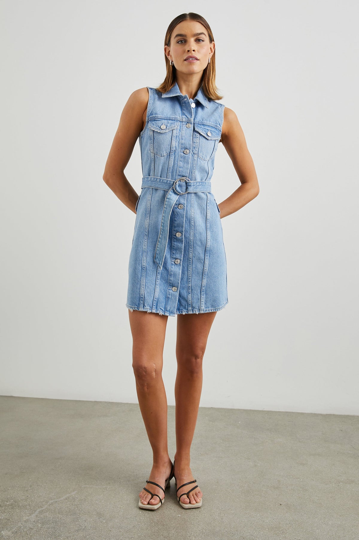 Wilshire Sleeveless Denim Mini Dress with a Full Button Front, Two Chest Button Flap Pockets, a Double Ring Belt, a Collar, and Front Vertical Seams in Blue Denim Colorway - Front Full Body View Featured on Model 