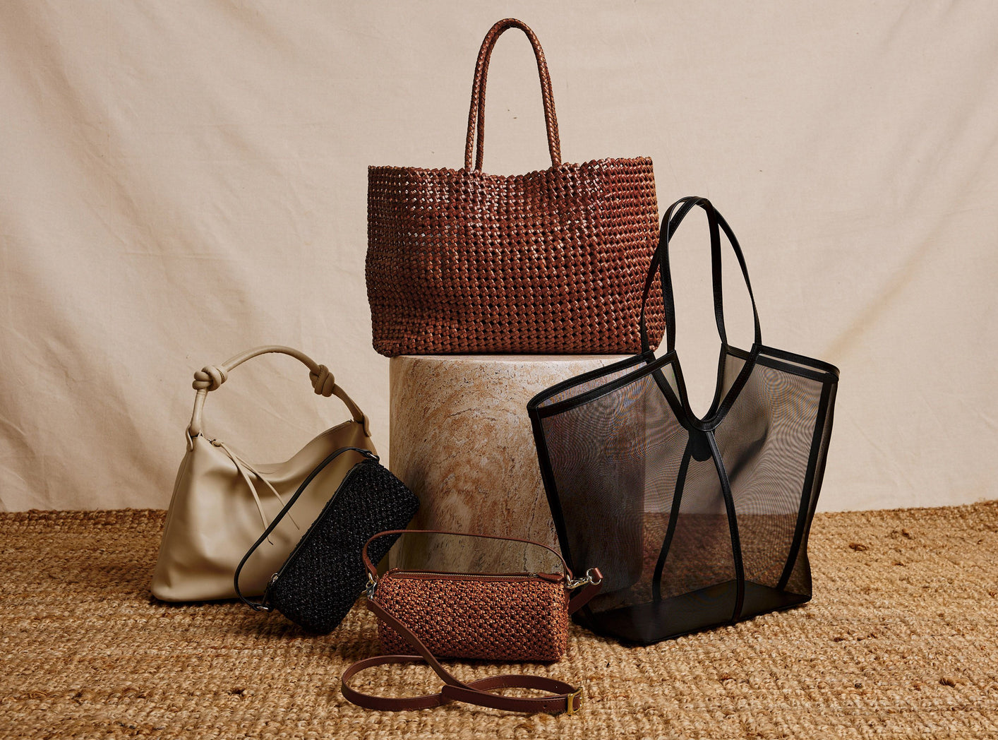 A styled product display of five handbags on a woven jute surface against a draped beige fabric backdrop. The collection includes a large brown woven tote bag, a black mesh tote bag, a cream leather shoulder bag, a black woven bag, a small brown woven pouch with a leather strap, and a small brown woven belt bag.