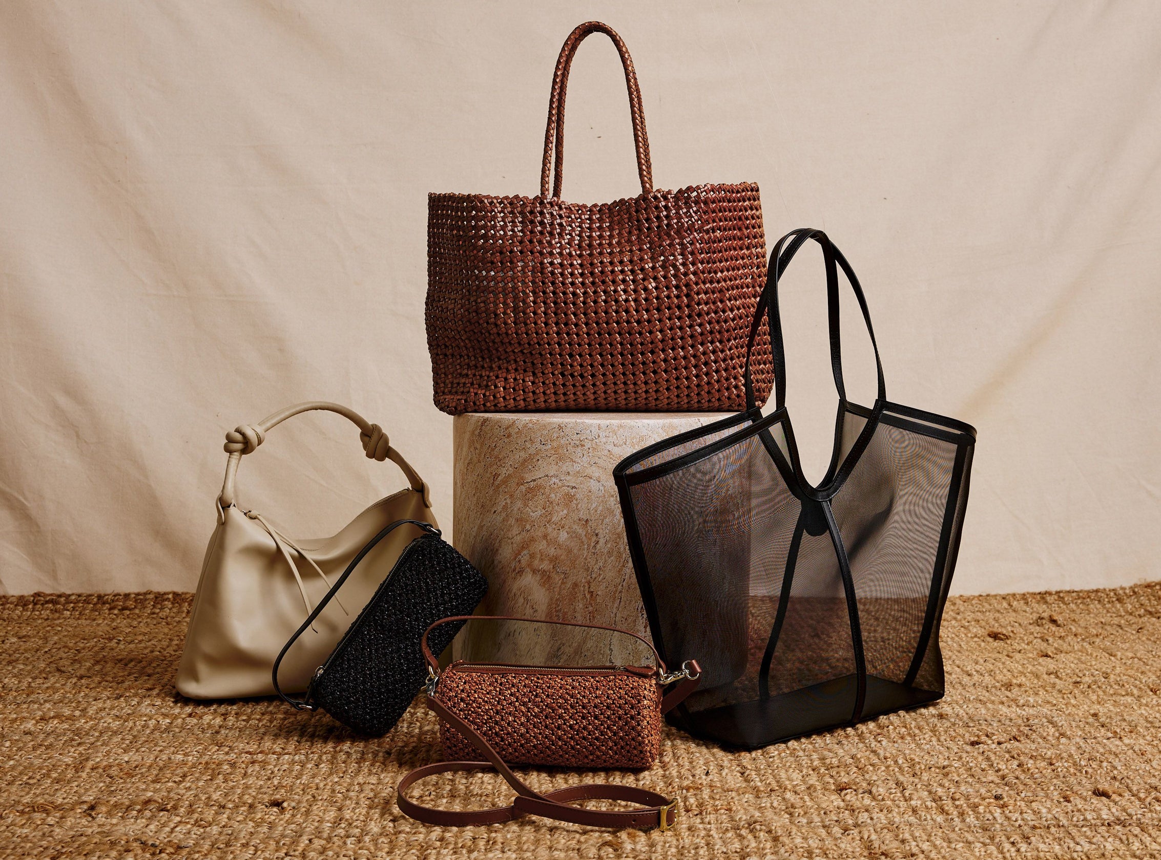 A styled product display of five handbags on a woven jute surface against a draped beige fabric backdrop. The collection includes a large brown woven tote bag, a black mesh tote bag, a cream leather shoulder bag, a black woven bag, a small brown woven pouch with a leather strap, and a small brown woven belt bag.