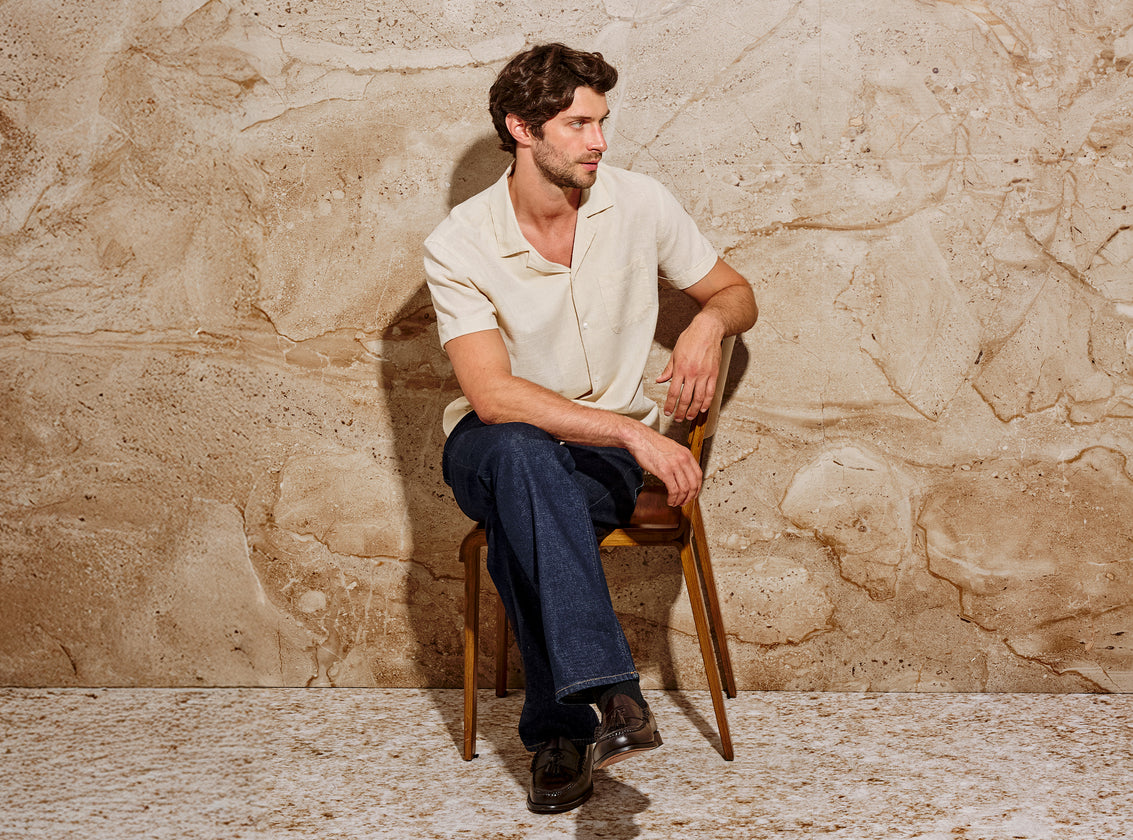 Man wearing short sleeve button down shirt sitting on a chair against a stone wall