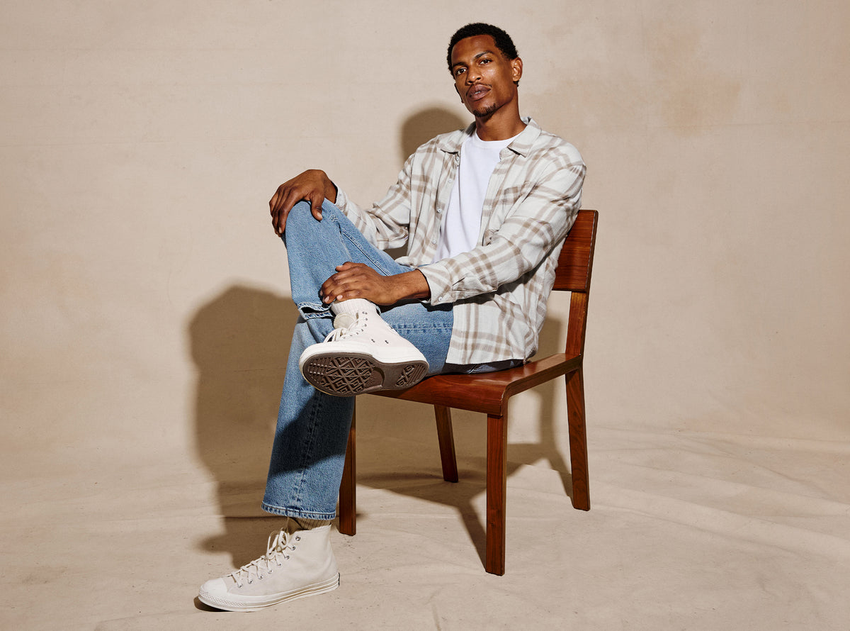 A man sits on a wooden chair wearing a beige and white plaid button-up shirt over a white t-shirt, blue jeans, and white high-top sneakers, set against a warm beige textured studio backdrop.