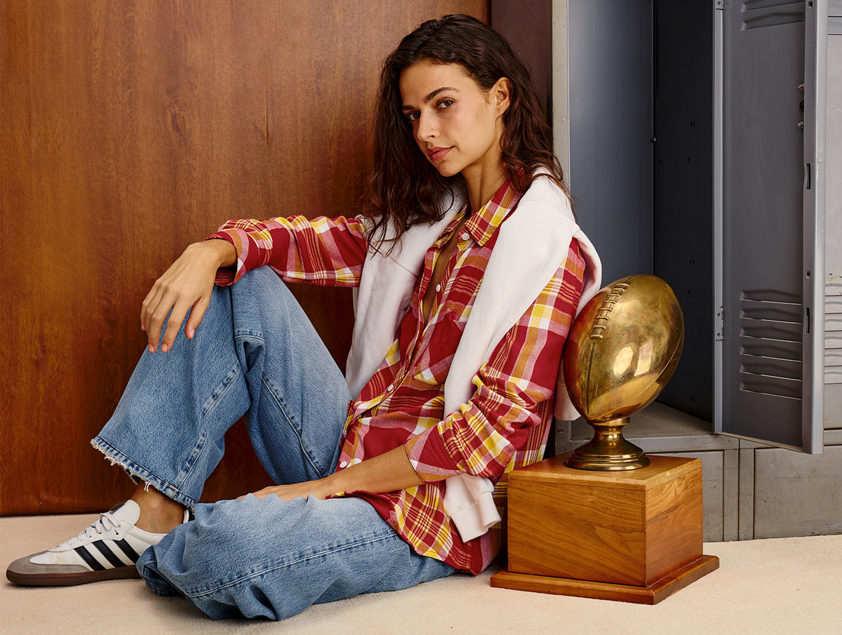 A woman sits on a floor in a locker-room-like setting, wearing a red and yellow plaid flannel shirt, blue jeans, a white zip-front vest, and white sneakers. A gold football trophy on a wooden base sits beside her.