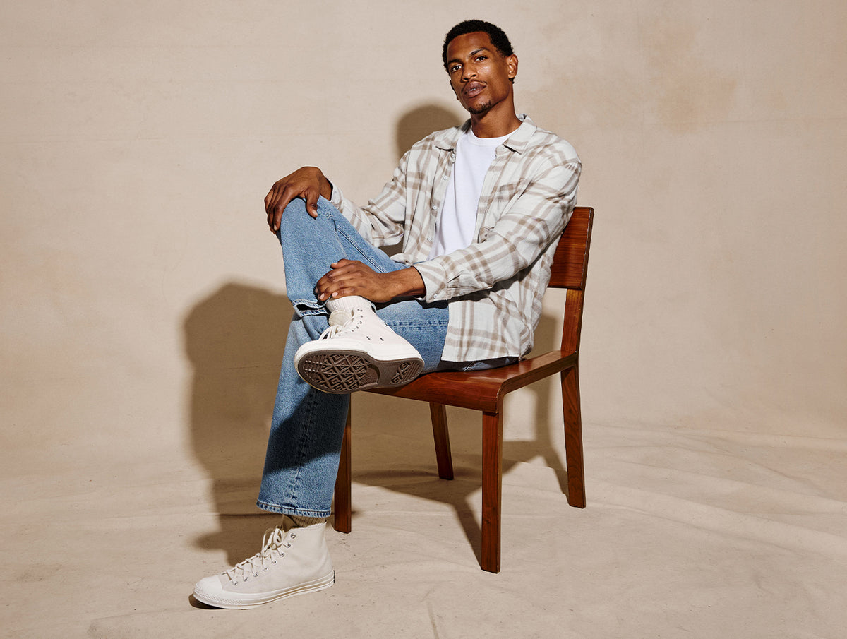 A man sits on a wooden chair wearing a beige and white plaid button-up shirt over a white t-shirt, blue jeans, and white high-top sneakers, set against a warm beige textured studio backdrop.