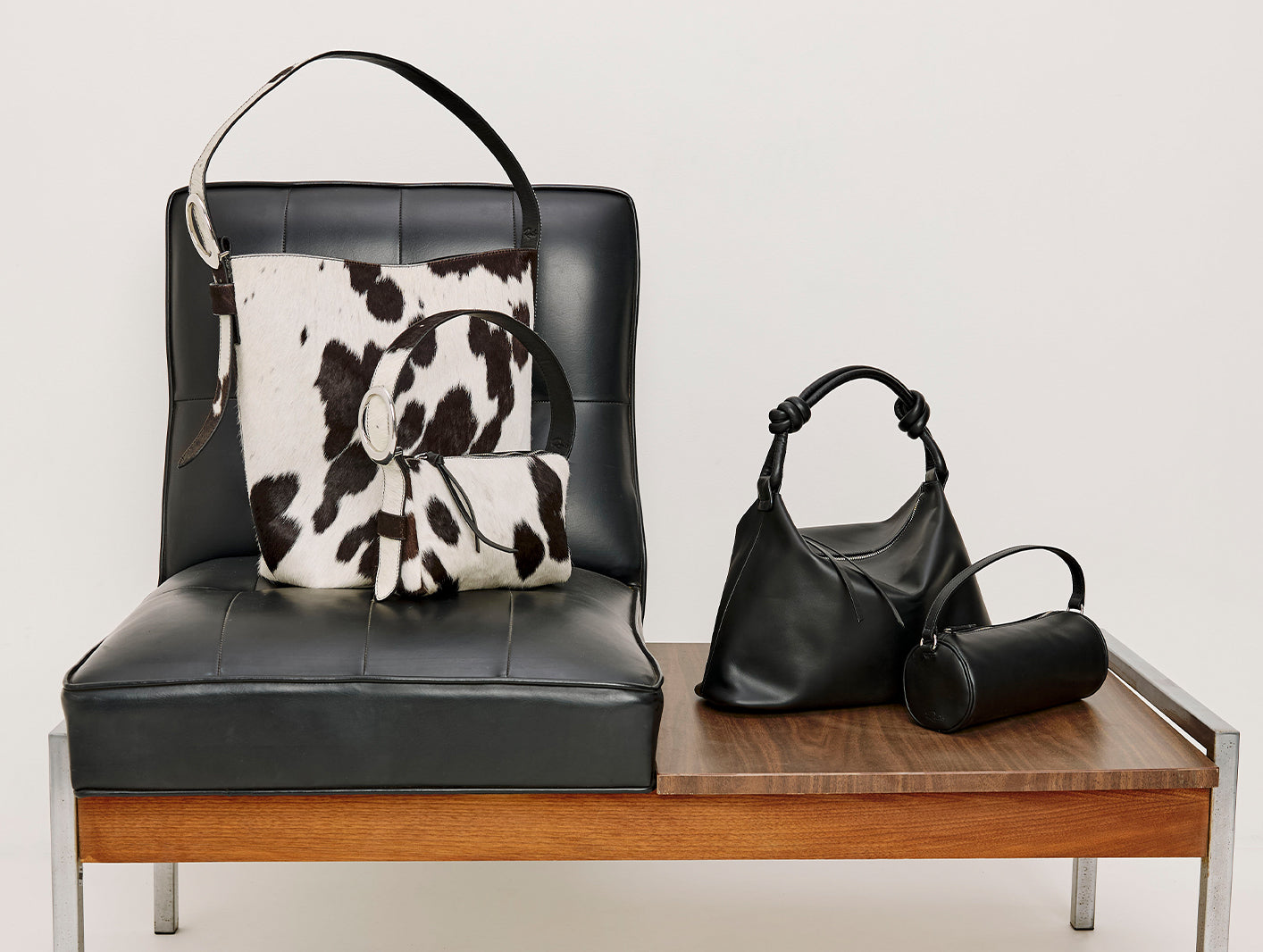 Two cow print handbags on black leather chair accompanied by two other black handbags on a table.