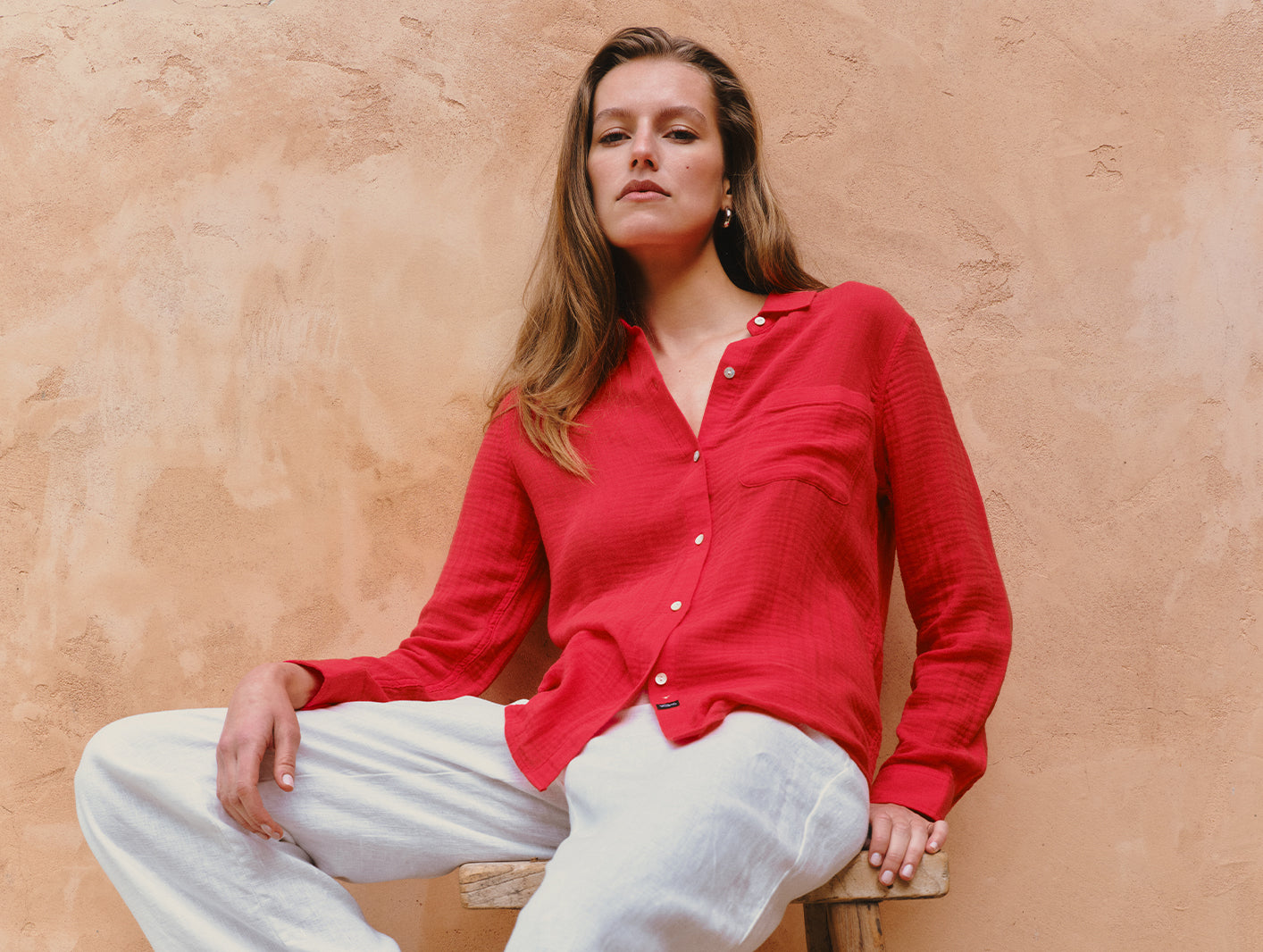 Woman wearing a red shirt and white pants sitting against a beige wall.