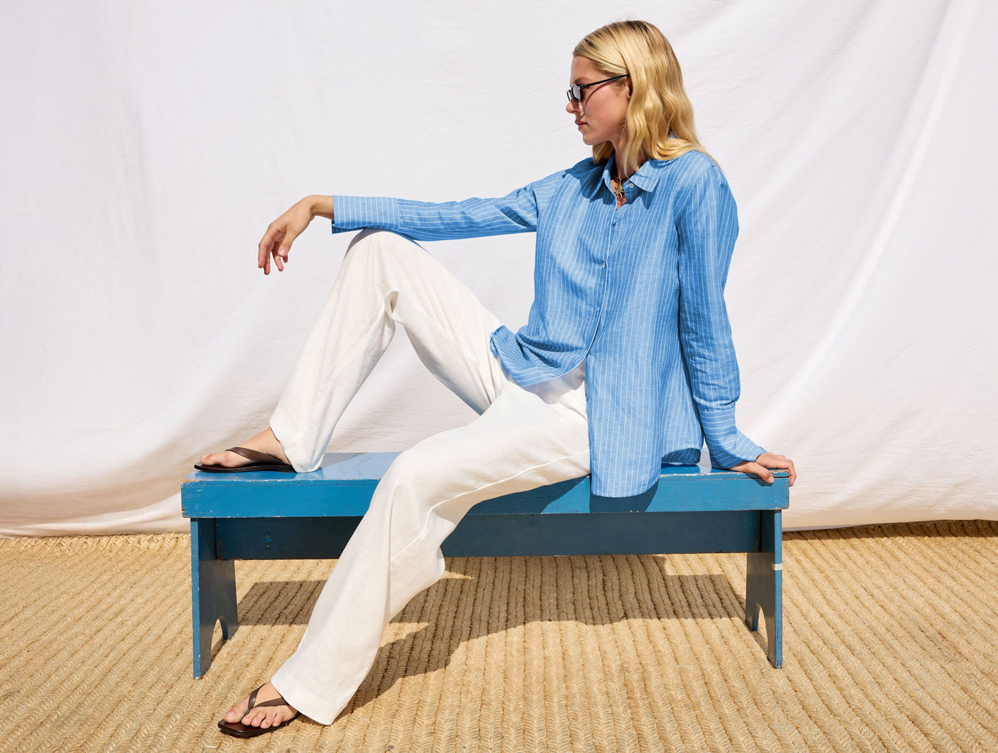 Woman sitting on a blue bench wearing a light blue shirt and white pants against a white background.