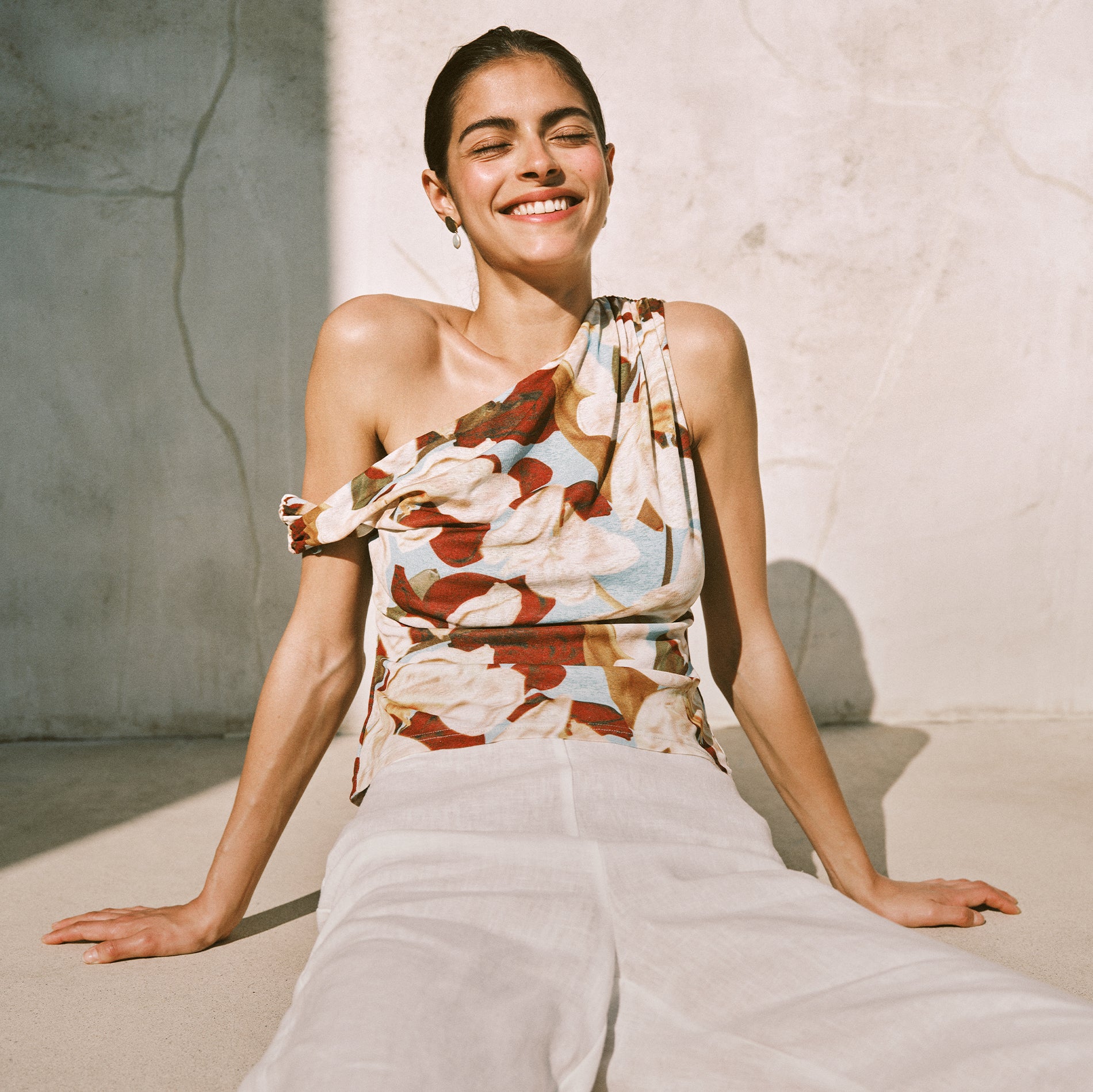 Woman wearing a floral top and white pants sitting against a light-colored wall.