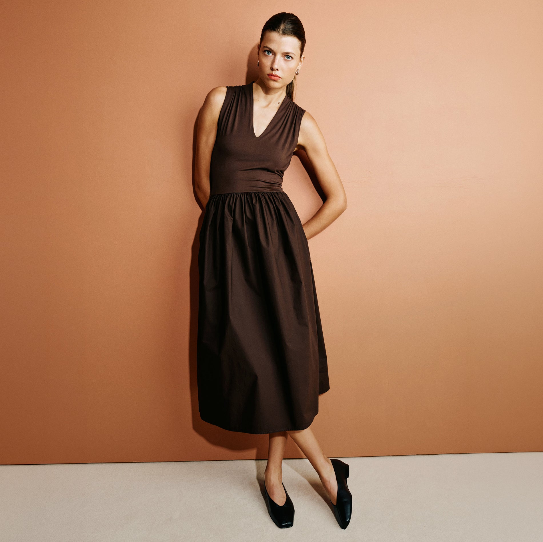 A woman modeling a sleeveless dark brown midi dress with a V-neckline and gathered waist, standing against a solid peach-colored studio backdrop.