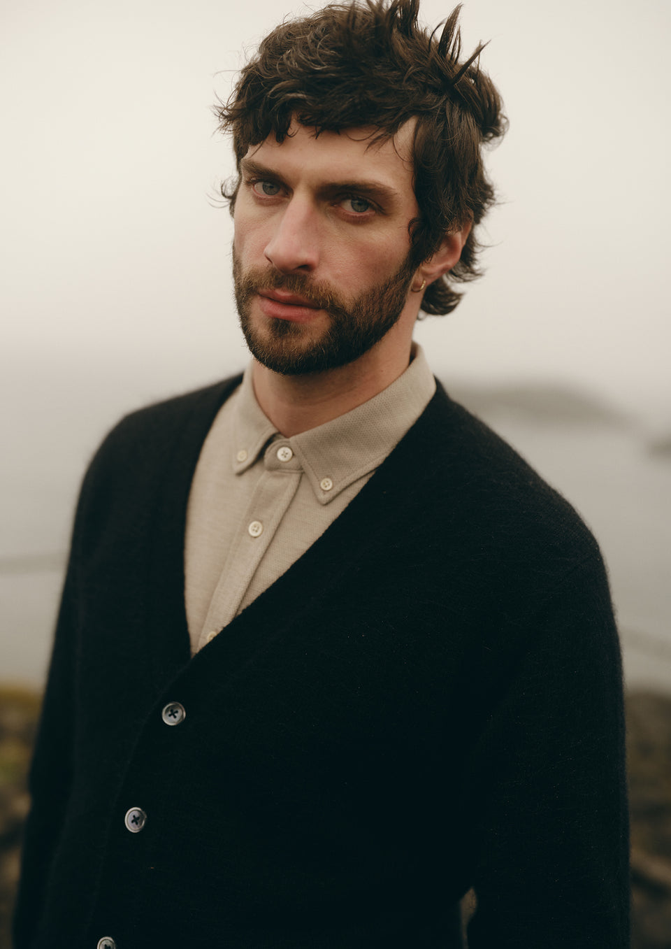 A man with dark curly hair and a beard wearing a black knit cardigan over a beige button-down shirt, photographed outdoors against a soft, misty background.