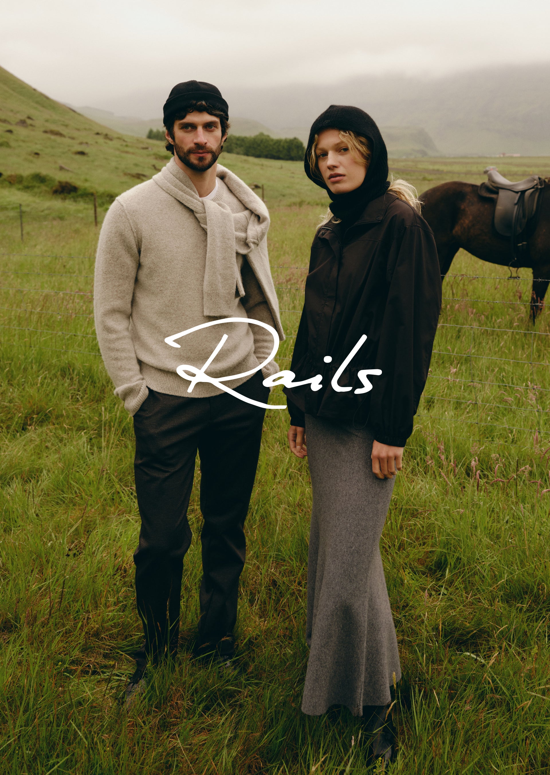 Two models standing in a grassy field with hills and a horse in the background, wearing dark and light neutral outerwear. A large white 'Rails' logo is overlaid across the center.
